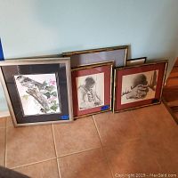 Photo showing multiple framed prints leaning against a wall on a tiled floor. Images include a bird on a branch, a person in thoughtful pose, and hands with head close-up.