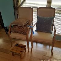 Two white arm chairs with wooden frames and light upholstery. Three accent pillows stacked on one chair: beige on top, brown shaggy textured below, blue scalloped pattern on the other chair. Small rustic wooden foot stool positioned in front of chairs.