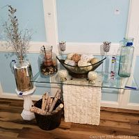 Photo of assorted vases, glass bowl with decorative woven spheres, metal basket with sticks, silver metal pitcher vase with dried stems, two glass votive holder candles, and tall glass bottles.