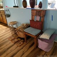 Photo showing wooden chair with blue seat cushion, red pillow on backrest, two baskets with pillows, four wooden framed sand art pictures on floor, wall mounted wine rack, round wooden side table.