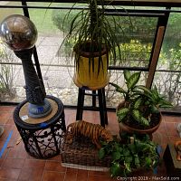 Overview of lot showing wicker table, pedestal with glass globe, black wooden plant stand holding yellow basket planter with tall plant, two terracotta pots with leafy plants, ceramic tiger figurine on wicker basket.