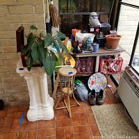 Wide view showing white column pedestal, bamboo plant stand, multiple plant pots, decorative owl statue, garden supplies on plant shelf, garden clock, and garden shoes.