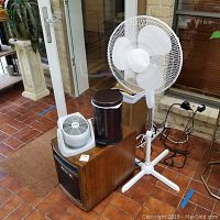 Four items arranged together: white standing fan, Duraflame heater with wood grain finish, dark brown pedal trash can, and white Holmes table top fan on top of heater