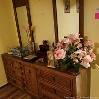 Wide view of wooden dresser top with multiple faux flower arrangements and assorted decorative items including clock and jewelry box