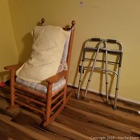 Wooden rocker with a cushioned seat, back cushion, and a pillow placed against beige wall with wooden floor