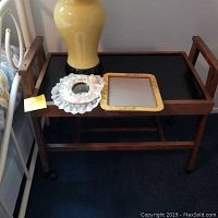 Photo showing wooden end table with black top, raised edges, side handles and wheels. There are decorative items placed on top including a yellow lamp, a lace doily and a small empty picture frame.