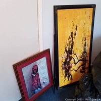 Two framed paintings leaning against a wall, one abstract yellow with black design, one portrait of child in red coat and hat.