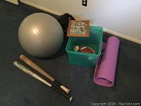 Wide shot showing yoga ball, two baseball bats, purple yoga mat, plastic bin containing football, baseball in plastic case, and framed signed Dallas Cowboys memorabilia, two black bags in background.