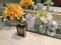 Photo showing the small Linden clock, orange and white faux flowers in vases, white plastic toothbrush holder, and glass tray on bathroom counter.
