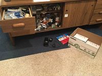 Wide angle photo showing open drawers with multiple small office supplies such as rubber bands, ink pads, rubber stamps, dispenser, and box with paper reams visible on blue carpeted floor.