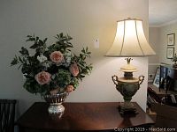 Photo showing the lamp and floral arrangement side by side on a wooden table, highlighting the lamp’s floral ceramic base and the floral arrangement's colors and composition.