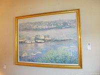 Framed coastal painting showing harbor with boats and town in the background, gold frame visible.