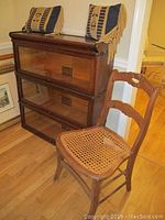 Front and side view of 3-shelf Globe Wernicke barrister bookcase with wooden chair with cane seat beside it