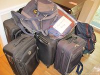 Overview of 6 rolling luggage bags including garment bags and totes stacked together in room corner