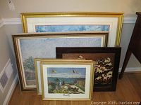 Lot of six framed prints and one mounted relief arranged stacked near a wall with visible corner of furniture and baseboard