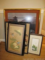 Photo showing grouped Asian artworks leaning against a wall, a visible mix of framed art including a large pagoda scene and smaller bird and flower themed paintings or needlepoints.