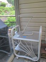 White rolling 2-shelf metal cart with tubular frame and angled supports, positioned near porch wall.