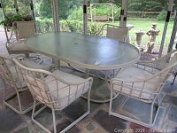 Wide view of the oval glass top table with all five chairs surrounding it on a stone patio indoors or in a sunroom.