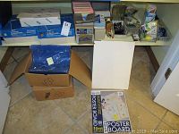 Boxes of office supplies including mailing envelopes, binder clips, highlighters, and white poster boards on the floor and shelves.