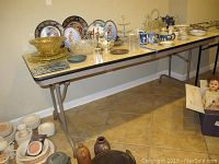 Folding table holding various collectible dishes and glassware, showing the full length on tiled floor.