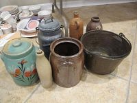 Multiple stoneware crocks and bottles, a green stoneware crock with floral design, a metal pail with lid and a copper bucket, arranged on the floor for display.