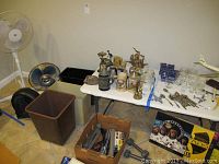 Shows three fans (white standing, black box, and silver desk) and three waste bins in front of folding table with various other items on table.