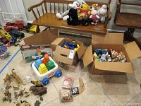 Overview photo of the lot showing a wooden bench with plush toys on it, boxes containing animal figurines and Little Tikes wagon with colorful block toys, and other scattered toys on the floor around the boxes.