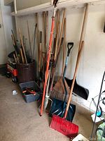 Wide angle photo of various yard tools lined up along a wall including long handled shovels, rakes, a red snow shovel, brooms, and bins with smaller tools and plant stakes.