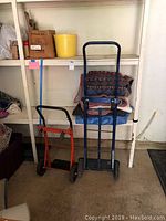 Photo of two hand trucks (one blue, one red/orange) placed on the floor near shelves with moving blankets stacked above them.