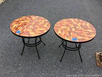 Full view of two round side tables with mosaic tile tops and black metal legs placed outdoors on pavement.