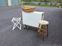Photo of white drop side crafting table, round wooden stool with padded seat, white folding luggage rack, and a wicker basket on top of the table, all on outdoor pavement surface.