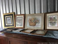 Several framed artworks and empty frames displayed on a dresser, including bird print, poem, flower basket, and cabin scene.
