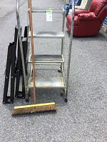 Full view of the metal step ladder with three worn steps and rubber wheels, adjacent to two black metal saw horses stacked together and a push broom leaning on the ladder.