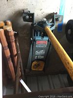 Side view of blue and yellow Lincoln 2 ton hydraulic floor jack leaning against a wall near wooden chair legs.