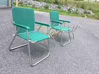 Two green folding chairs with metal tubular silver frames and green vinyl seats and backs. Accompanied by two brass racks with curved elements, placed on pavement outside.