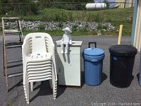 Shows 8 stacked white plastic patio chairs, wooden painters ladder, white storage cabinet, two large plastic trash cans in blue and black, and a decorative doll figure sitting on the cabinet.