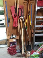 Full view of lot showing a collection of long-handled yard tools including shovels, pick axe, loppers, and electrical corded Toro leaf blower packed in a box with an extension cord on floor.