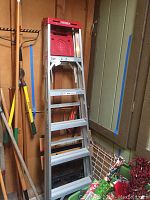Aluminum Werner 6ft step ladder leaning against wall with rakes and tools near it; ladder has 6 steps with red plastic top and made in USA label