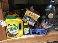 View of Preen Weed Preventer container, Miracle-Gro plant food, solar lights box, mole killer container, and blue basket with garden accessories on wooden shelf.