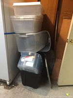 Stack of plastic storage bins in corner near a fridge and a door, includes clear/translucent and black bins, some with white lids.