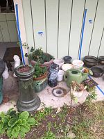 Showing multiple garden planters of various sizes and materials including a large terracotta-colored planter, white and dark ceramic or plastic pots, a ceramic owl figurine, green plastic watering can, and assorted plants in some pots.