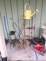 View of 3 metal plant stands and yellow watering can, placed outside near a wall showing rust and wear.