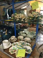 View of the three Capidimonte floral figurines on a glass shelf with ceramic cups and a serving dish below