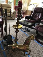Photo showing full view of brass candlestick, brass horse statue, and fireplace tools rack with tools in living room environment.