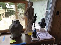 Wide shot showing all five items: alabaster statue, bronze statuette, black bust sculpture, alabaster vase, and decorative plaque on table near window.