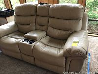 Full view of beige leather dual recliner loveseat showing padded seats, backrests, armrests, and center console with cup holders.