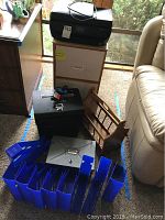 Shows HP Office Jet 4652 printer on top of a beige file cabinet with drawers, black metal lockable safe/box in front, wooden magazine rack beside, and multiple blue plastic file organizers spread out in front.