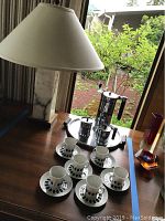 Photo of full set showing marble table lamp, Sunbeam coffee pot, creamer, sugar set on tray, and six German tea cups with saucers on wooden table by window.