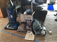 Group shot showing all three appliances: Hoover WindTunnel vacuum, Dirt Devil steamer, Black and Decker vacuum with box, charger and manual on carpeted floor next to furniture.