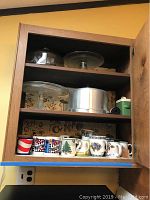 Shelf with two footed clear glass cake plates, metal cake cover, glass lid, and multiple coffee mugs